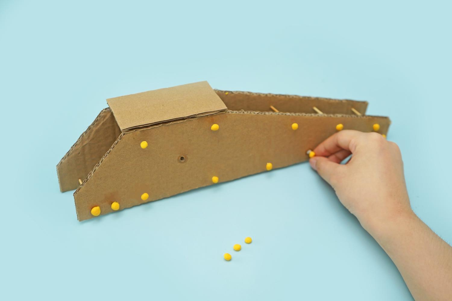 A hand adds small yellow modelling clay balls to a cardboard crane structure, already fitted with toothpicks, on a light blue surface.