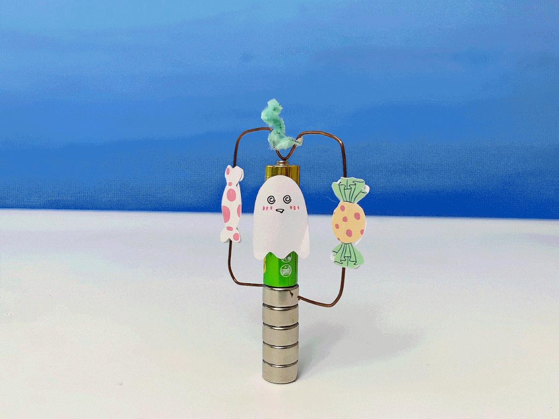 A small, completed spinning ghost science model, featuring a ghost and sweets attached to a wire frame around a battery and magnet, sits on a white surface against a blue background.