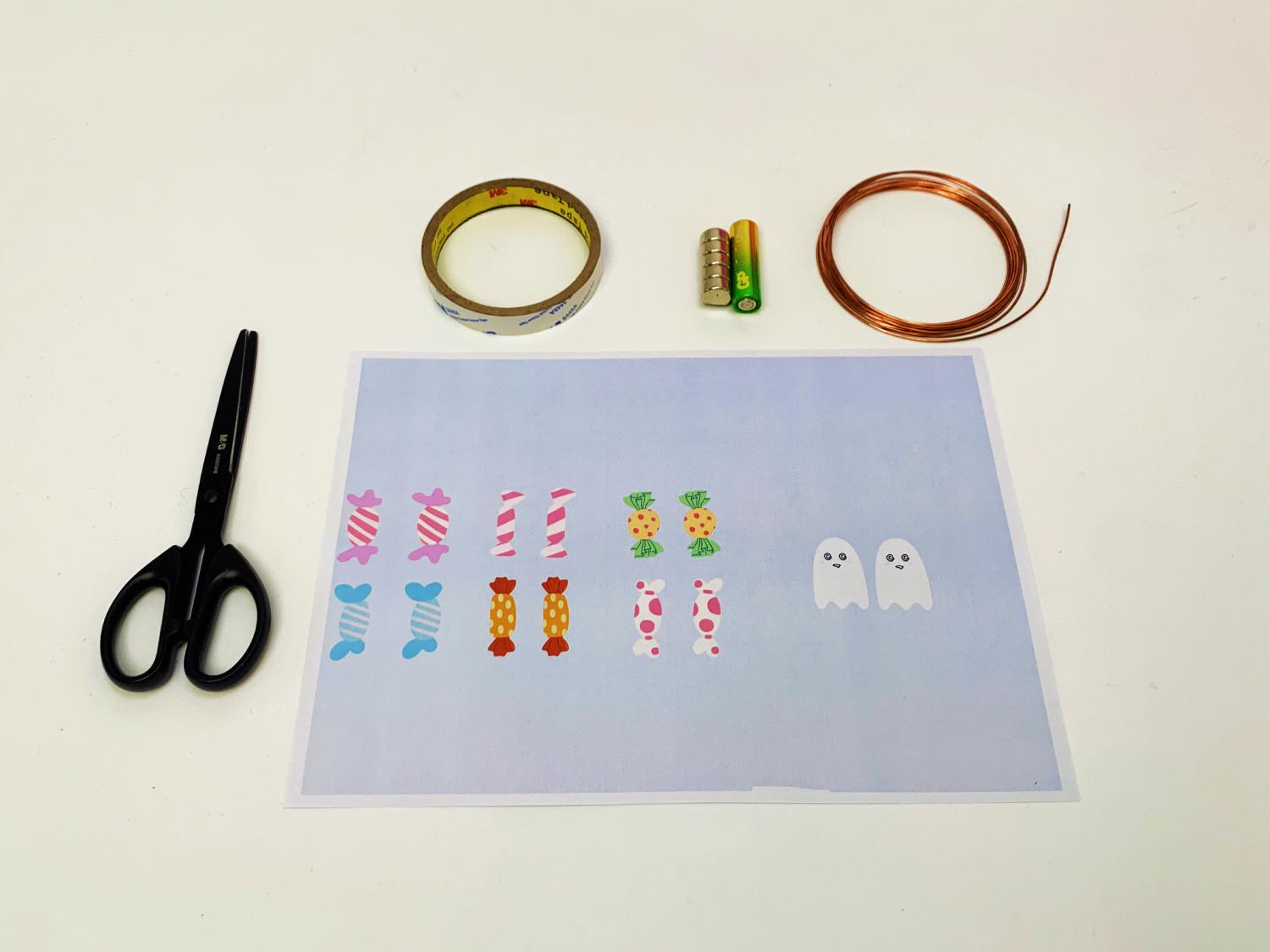 A pair of scissors, a roll of tape, a small battery, a length of copper wire, and a sheet of paper with drawn sweets and ghosts are arranged on a white surface, ready for a craft project.