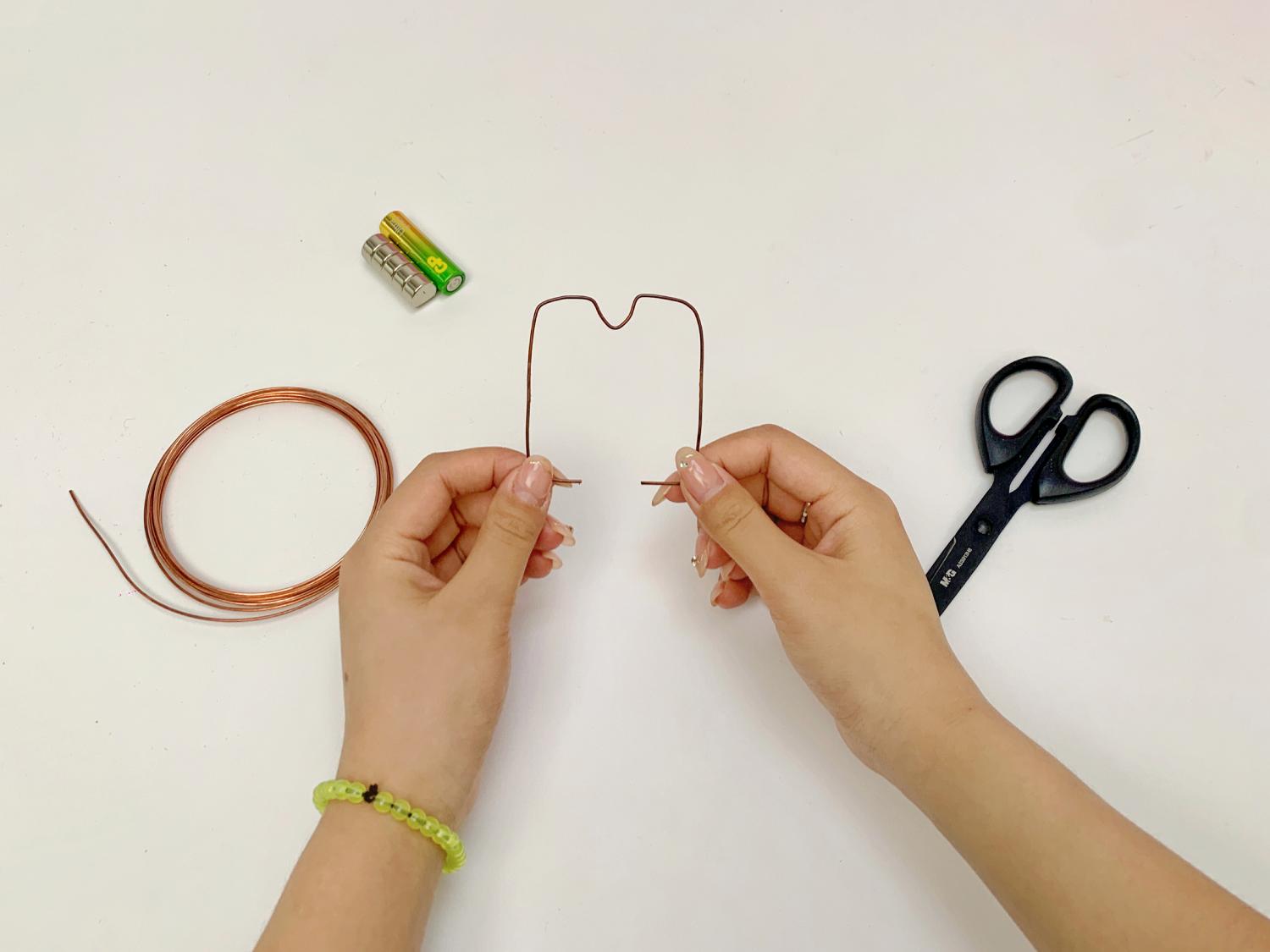 Two hands carefully shape a piece of copper wire into a small frame, with other craft materials including batteries and scissors nearby on a white surface.