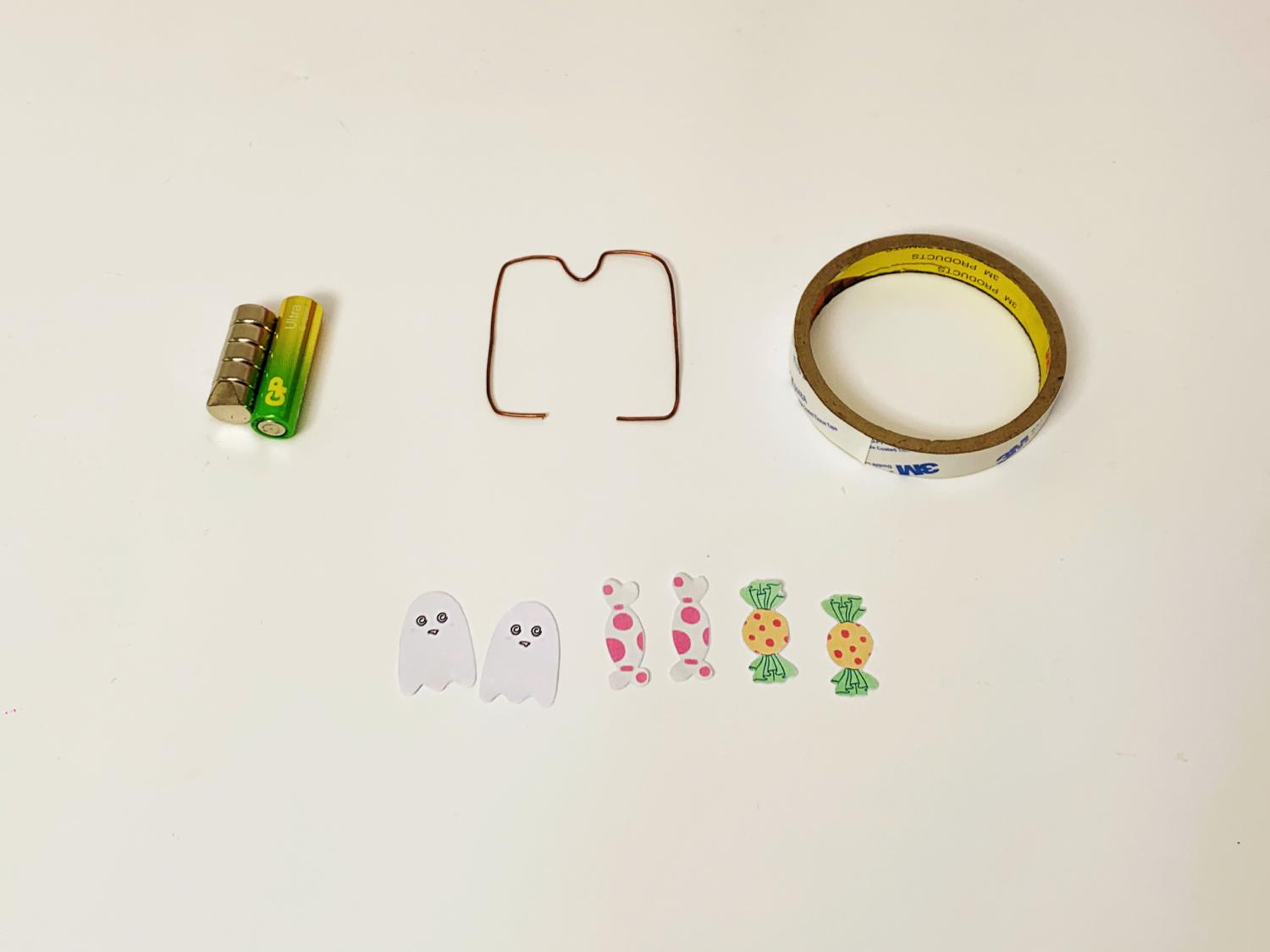 A battery, a small magnet, a length of copper wire shaped into a rectangle, a roll of sticky tape, and some paper cut-outs of ghosts and sweets are laid out on a white surface.