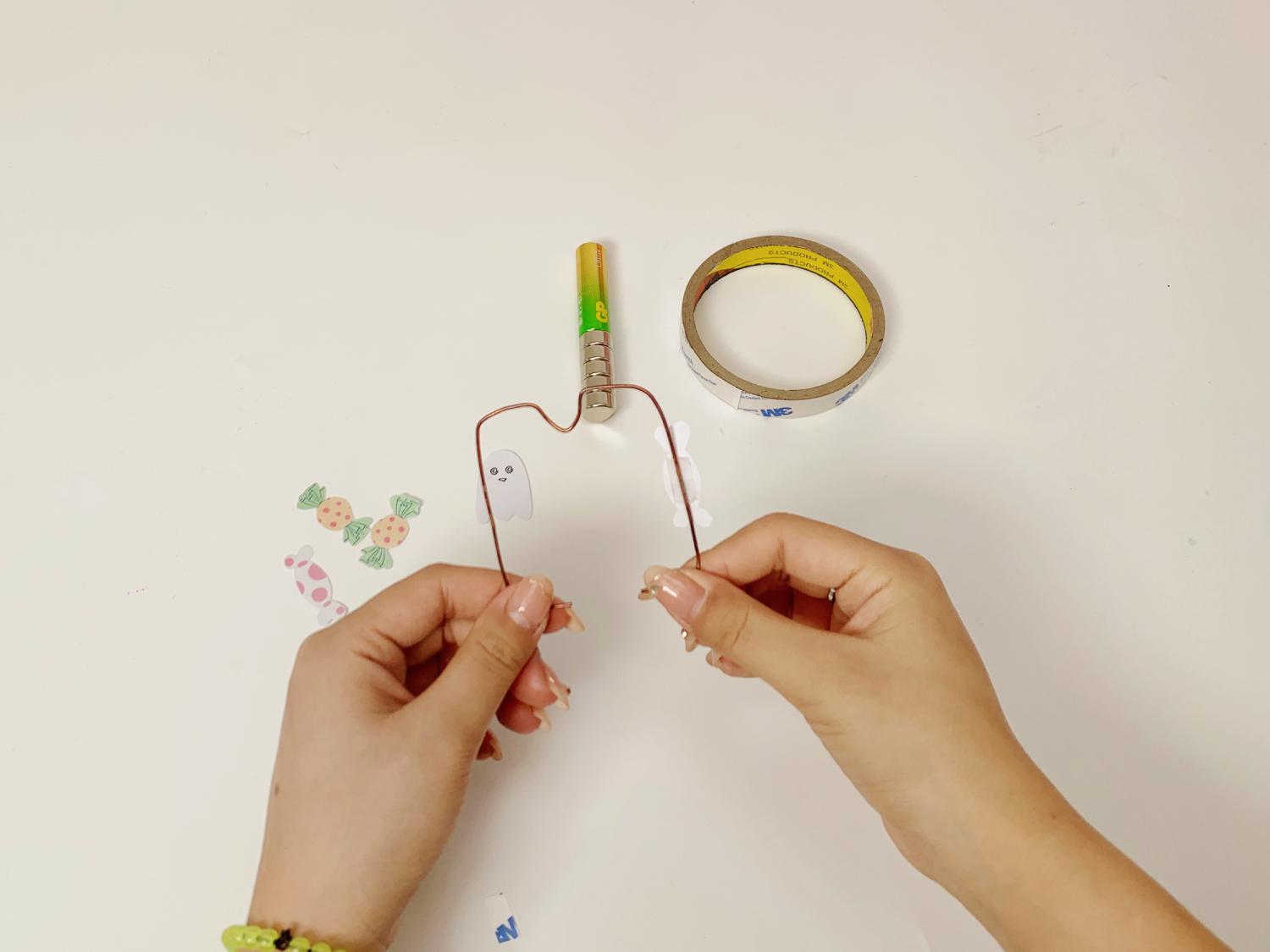 Two hands hold a small copper wire frame, with a ghost cut-out attached, whilst a battery, magnet, and tape sit nearby on a white surface.