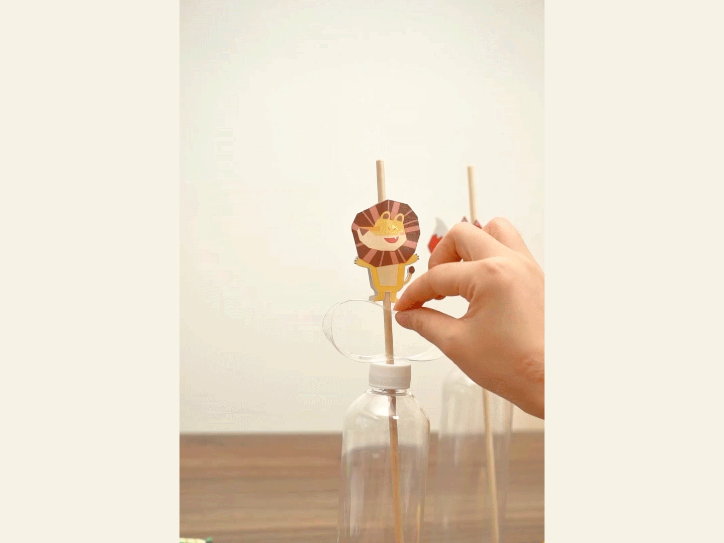 A hand attaches a paper lion cutout to a wooden stick inserted into a clear plastic bottle, likely part of a DIY craft project.