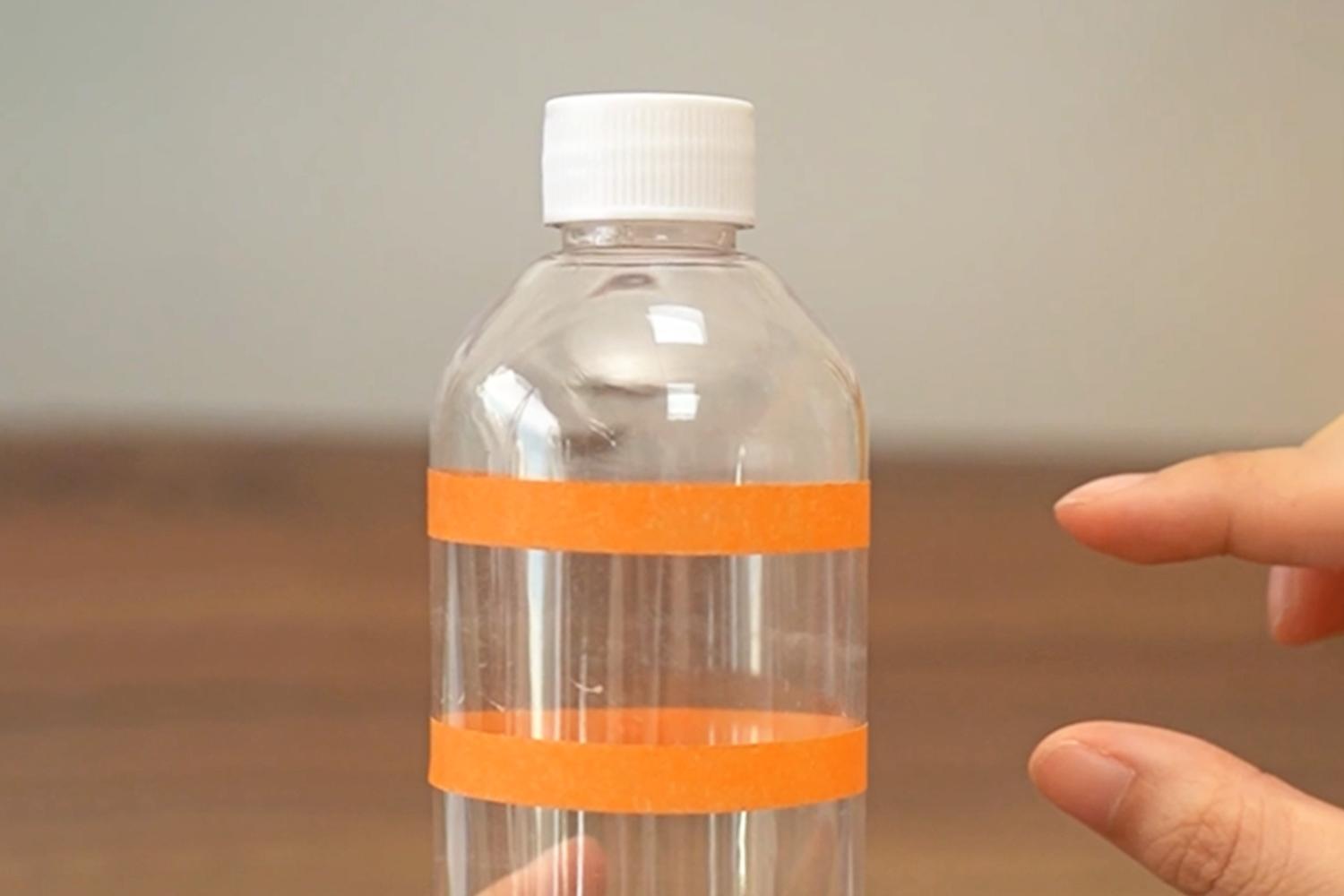 A clear plastic bottle, with two bands of orange tape around it, is held in one hand, whilst another hand gestures nearby, on a wooden surface.