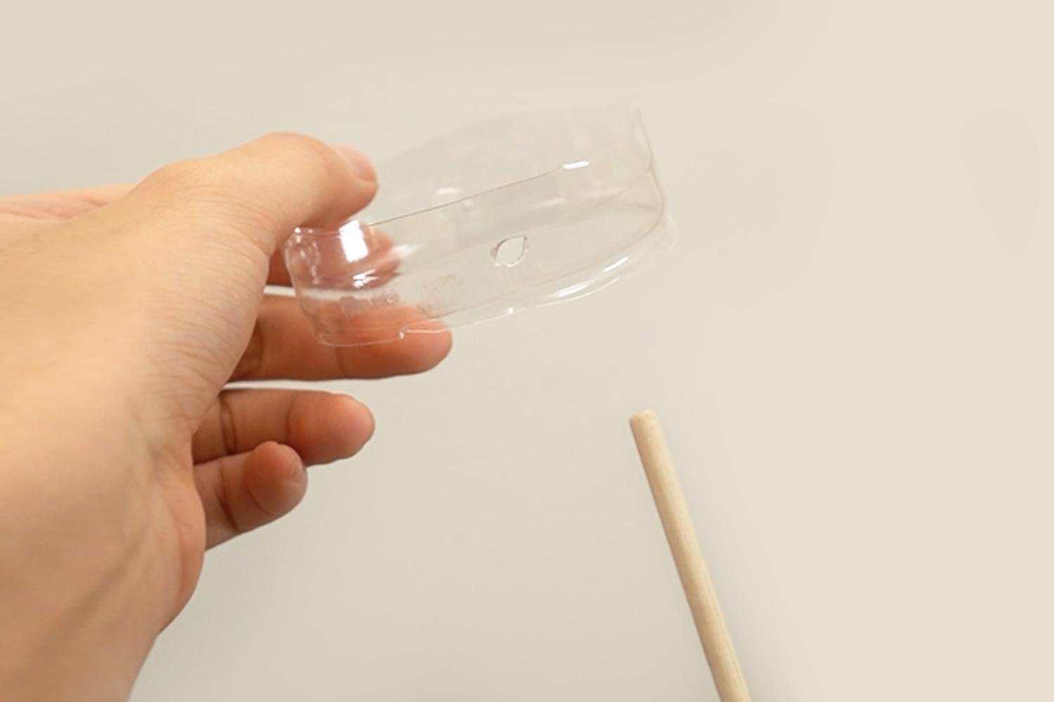 A hand holds a clear plastic bottle top with a hole in it above a small wooden dowel on a white surface.