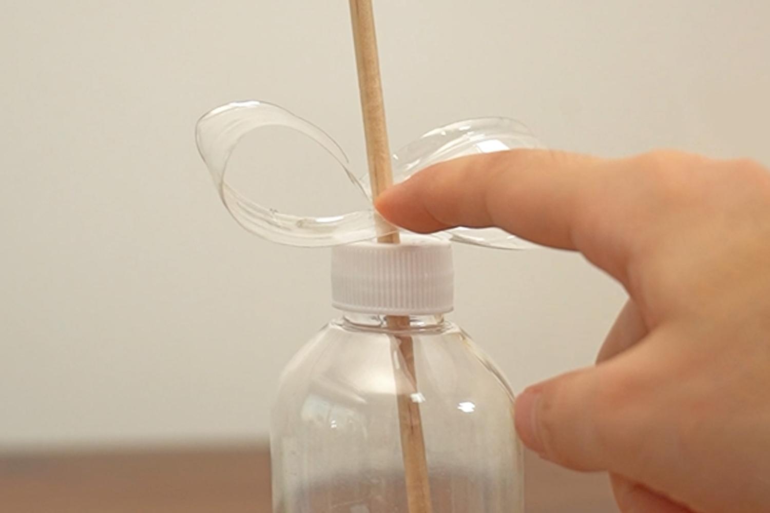 A hand carefully positions a cut-off plastic bottle top around a wooden dowel, which is inserted into the bottle's cap.