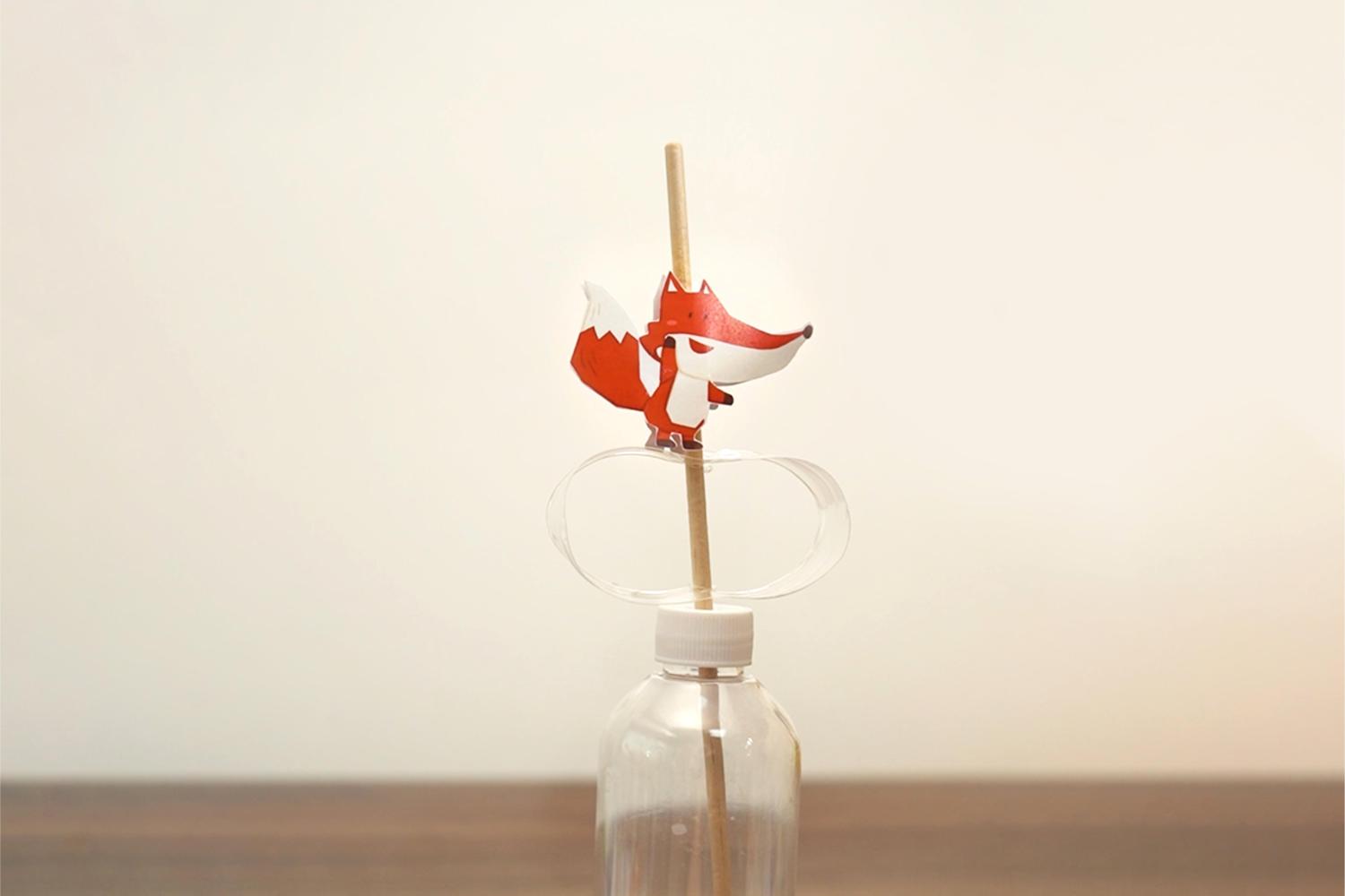 A small, illustrated fox sits atop a wooden stick which is pushed through a plastic bottle top and into a clear plastic bottle, on a wooden surface.