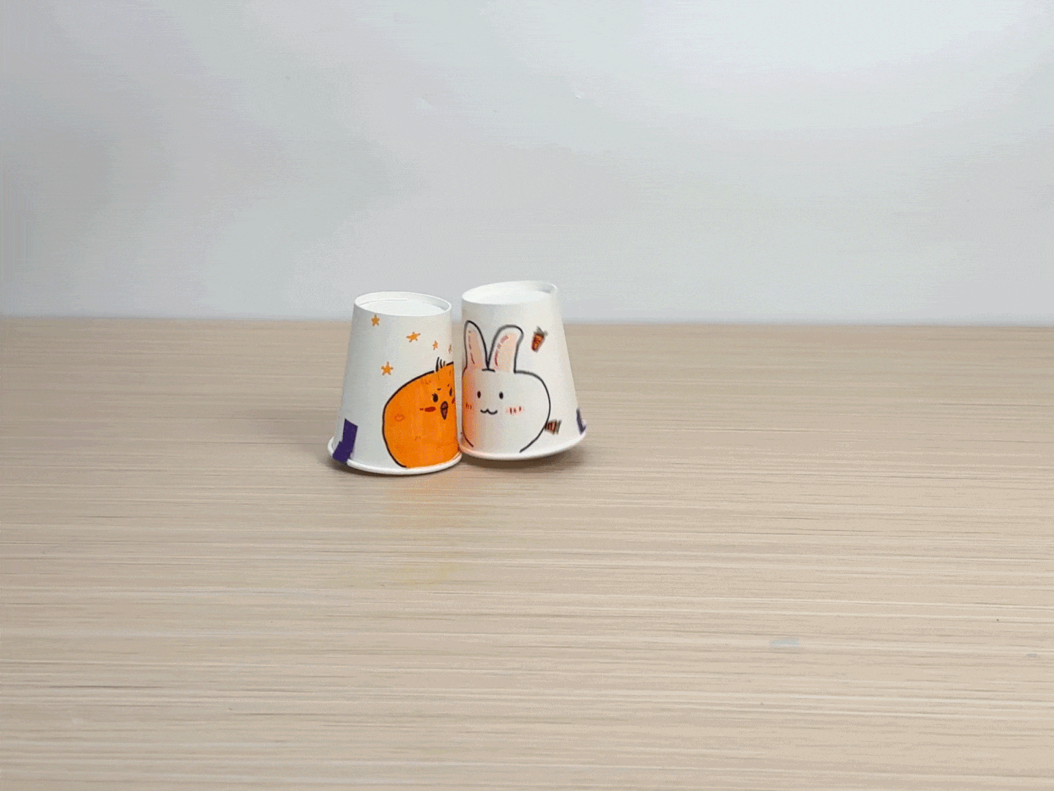 Two decorated paper cups spin on a wooden table. One cup features a cartoon chick and stars, while the other has a cartoon rabbit.