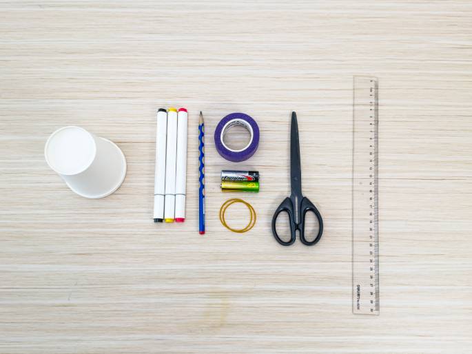 An assortment of craft supplies lies on a light wood-grain table, arranged for a project. The items include a white paper cup, three white markers, a pencil, a roll of purple tape, two AA batteries, a rubber band, a pair of scissors, and a clear plastic ruler. This flat lay image showcases the materials needed for a simple DIY activity, likely a science experiment or children's craft.
