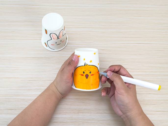 Two hands are decorating paper cups on a light wood-grain surface. One cup, lying on its side, already has a drawing of a bunny and carrots. The other cup, held upright by one hand, features a drawing of a chick and stars. The other hand is using an orange marker to add details to the chick's cheek. This close-up image captures the creative process of decorating paper cups, likely for a children's craft activity.