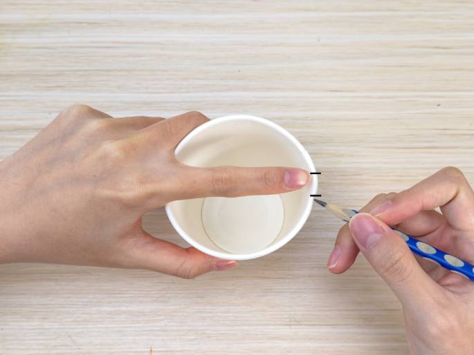 Two hands are shown working with a white paper cup on a light wood-grain surface. One hand holds the cup steady while the other hand uses a pencil to make small marks on the rim. The index finger of the hand holding the cup is placed on the rim, seemingly measuring or marking a specific distance. This close-up image suggests a DIY or craft activity involving precise marking on a paper cup.