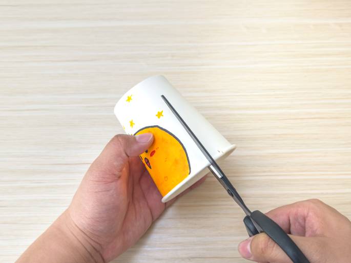 Two hands are shown manipulating a decorated paper cup on a light wood-grain table. One hand holds the cup, which features a drawing of a chick and stars. The other hand uses scissors to cut into the top edge of the cup. This image illustrates a step in a craft project, likely involving modifying the paper cup for a specific purpose.