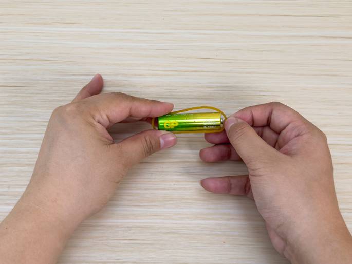 Two hands are manipulating a AA battery and a rubber band against a light wood-grain background. One hand holds the battery while the other hand pulls a rubber band that is looped around one end of the battery. This close-up image suggests the process of attaching the rubber band to the battery for a science experiment or a small DIY project.