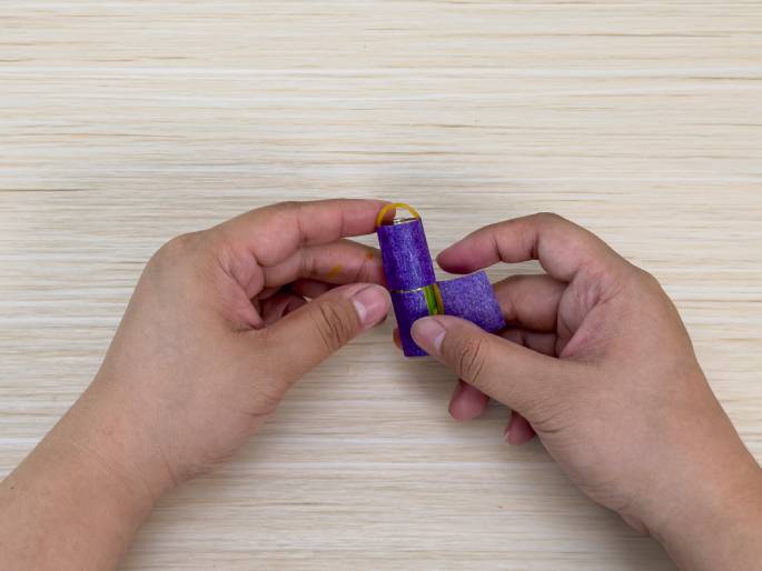 Two hands are working with a AA battery, a rubber band, and purple tape on a light wood-grain surface. A rubber band is looped around one end of the battery, and purple tape is being wrapped around the battery. One hand holds the battery and the beginning of the tape, while the other hand secures the tape in place. This close-up image suggests a step in a DIY project or science experiment involving assembling a small device or mechanism.