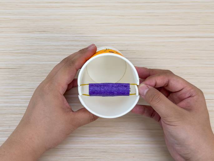 Two hands hold a white paper cup on a light wood-grain surface. A battery wrapped in purple tape is secured to the sides of the cup with rubber bands, creating a handle-like structure across the opening. The cup appears to be part of a DIY project or craft, possibly a homemade toy or device.