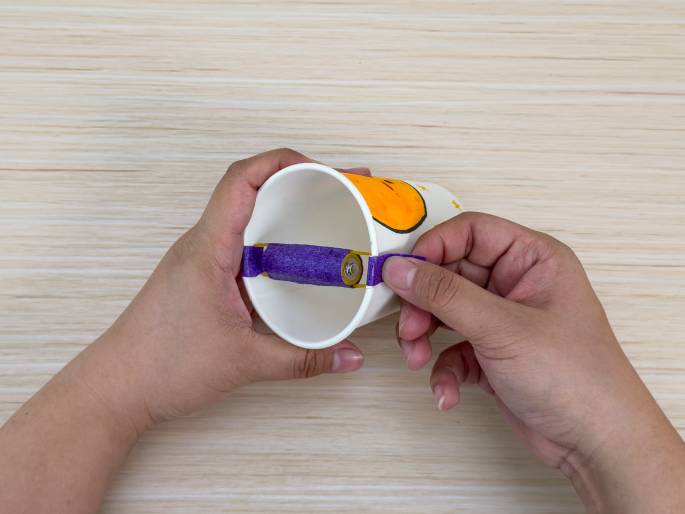 Two hands are holding a decorated paper cup on a light wood-grain surface. The cup has a drawing of a chick and stars. A battery, wrapped in purple tape, is attached to the cup with rubber bands, forming a handle. One hand holds the cup while the other adjusts the purple tape. This image likely depicts a stage in a children's craft project.
