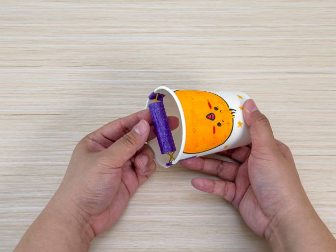 Two hands are manipulating a decorated paper cup with a handle on a light wood-grain surface. The cup features a drawing of a chick and stars. The handle is made from a battery wrapped in purple tape and attached to the cup with rubber bands. The hands are holding the cup at an angle, showcasing the handle. This image likely illustrates a step in creating a simple craft or toy.