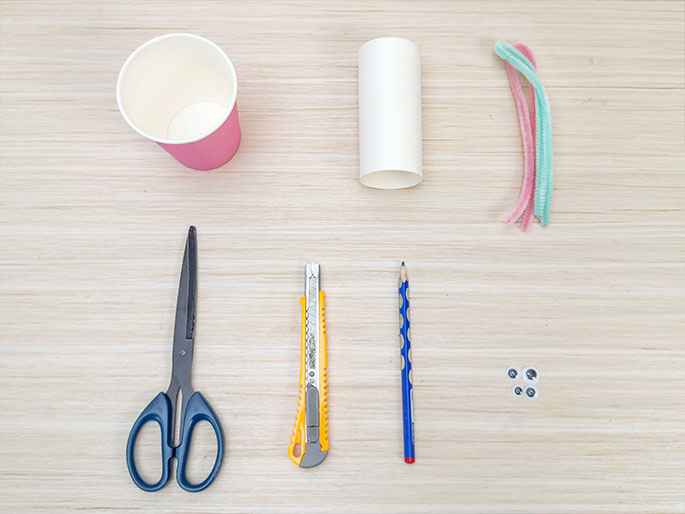 An overhead flatlay image displays various craft supplies arranged on a light woodgrain surface. The items include a pink paper cup, a cardboard tube, a pair of blue-handled scissors, a yellow utility knife, a blue pencil, a few pastel-colored pipe cleaners, and a small cluster of googly eyes. These materials appear to be gathered for a children's craft project.