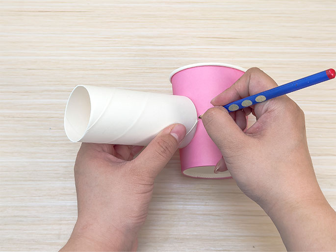 Two hands are shown in the process of making a craft project. One hand holds a white cardboard tube against a pink paper cup, while the other hand uses a blue pencil to trace the end of the tube onto the side of the cup. The hands and materials are positioned on a light woodgrain surface. This image captures a step-by-step crafting process, likely aimed at children.