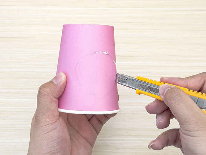 Two hands are shown working with a pink paper cup on a light woodgrain background. One hand holds the cup while the other hand uses a yellow utility knife to carefully cut out a circle that has been traced onto the side of the cup. The process suggests a crafting activity, and the precision required indicates it's likely a demonstration for older children or adults.