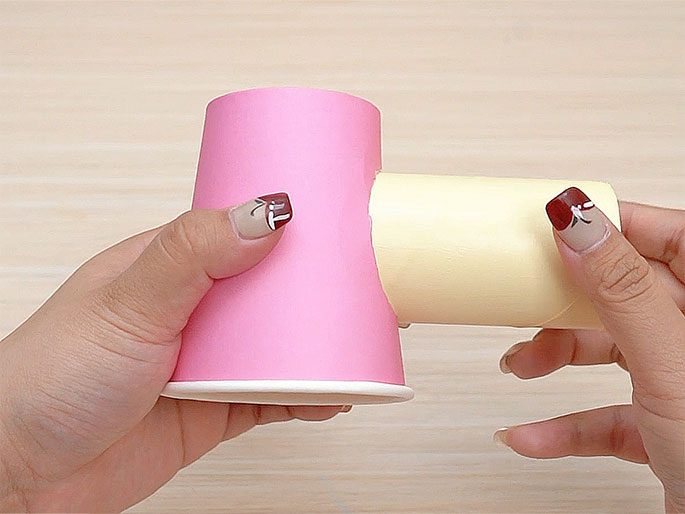 Two hands hold a pink paper cup with a cardboard tube inserted into a circular opening cut into its side. The tube extends horizontally from the cup. The background is a light woodgrain surface. The fingernails on the hands holding the craft project have subtle nail art. The image showcases a simple construction made from everyday materials, likely part of a children's craft activity.