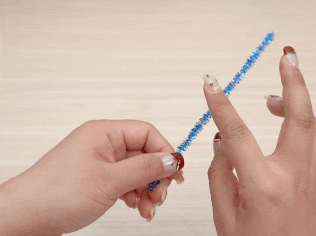 Two hands with decorated fingernails hold a sparkly blue pipe cleaner against a plain, light-colored background, likely a wooden table or desk. The hands manipulate the pipe cleaner, bending it and shaping it, presumably as part of a craft or DIY project.