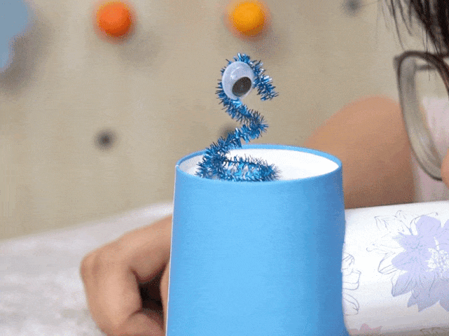 A close-up view of a light blue paper cup. A shiny blue pipe cleaner is inserted into the cup, and a googly eye is attached to the top, creating a small puppet. A person wearing glasses is partially visible in the background. The background features a light-colored wall with blurred orange and yellow circles. This video demonstrates a simple craft project, likely aimed at children.