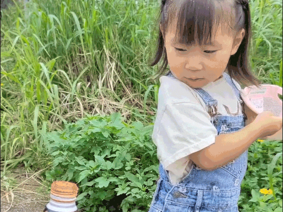 A small child in denim overalls plays with a modified drink bottle that sprays water like a fountain.