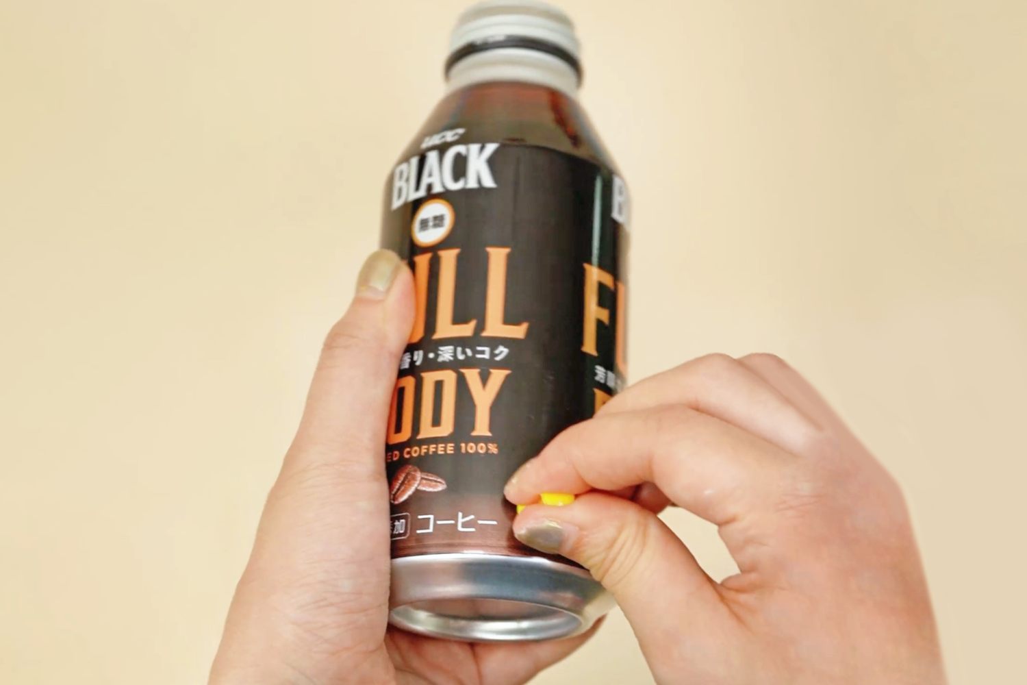 Two hands hold a can of Boss Black Full Body coffee, with one hand placing a small yellow object on the can's label, against a pale beige background.