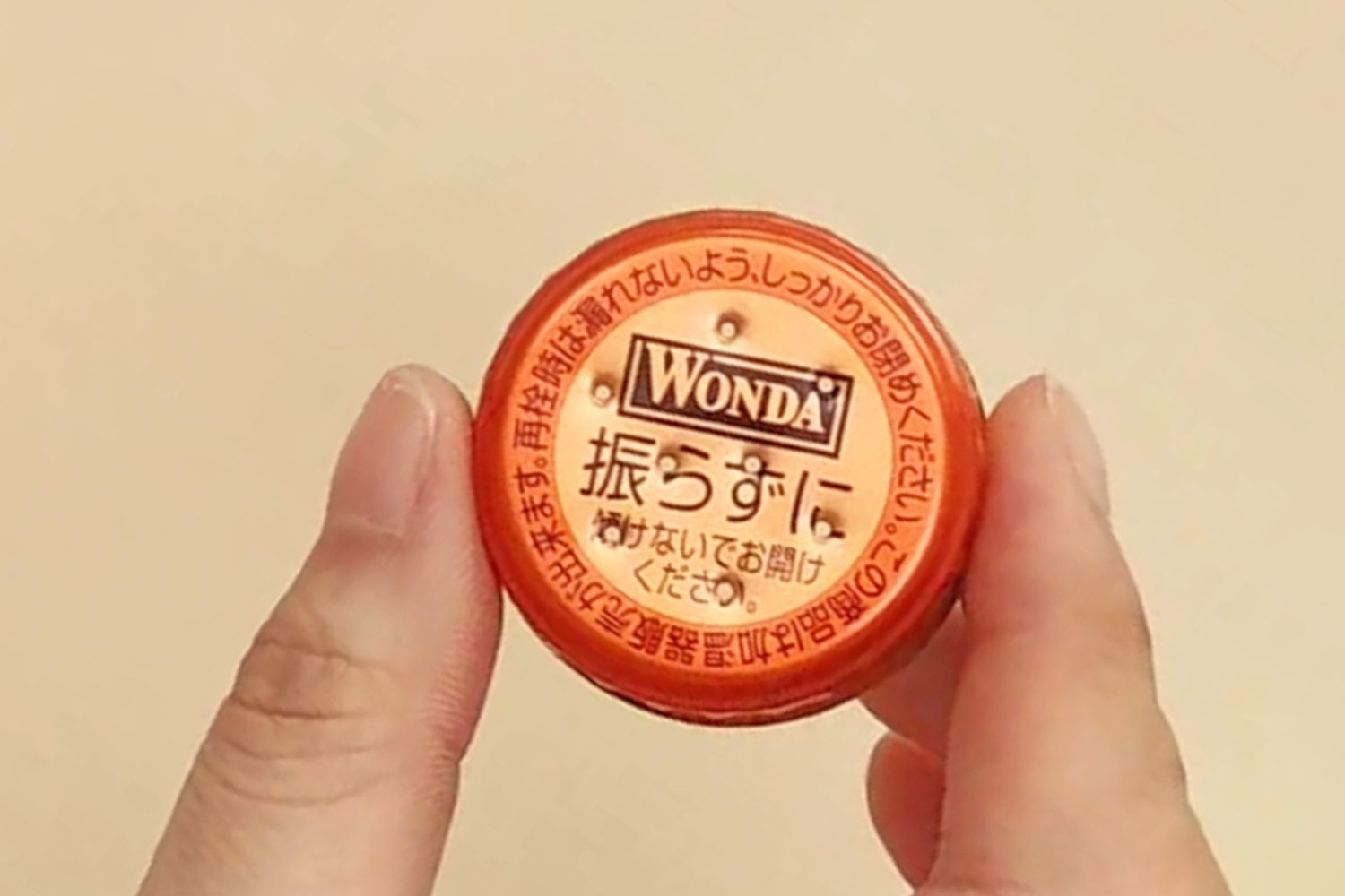 A finger and thumb gently hold a small, round, orange lid with Japanese writing and the word Wonda on it, against a pale background.