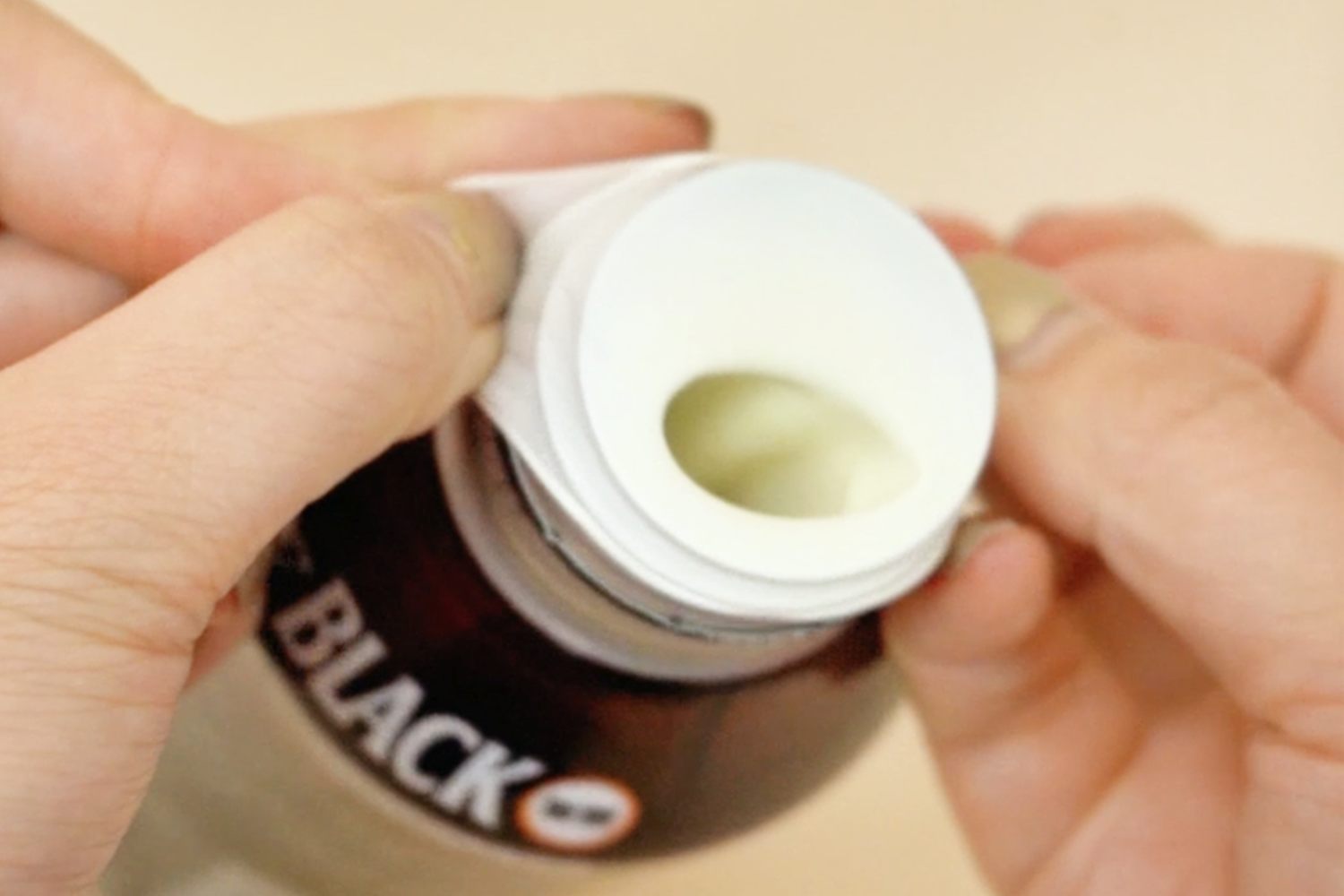 Two hands carefully remove a white plastic lid from a dark-coloured bottle, revealing a pale yellow substance beneath, against a pale background.