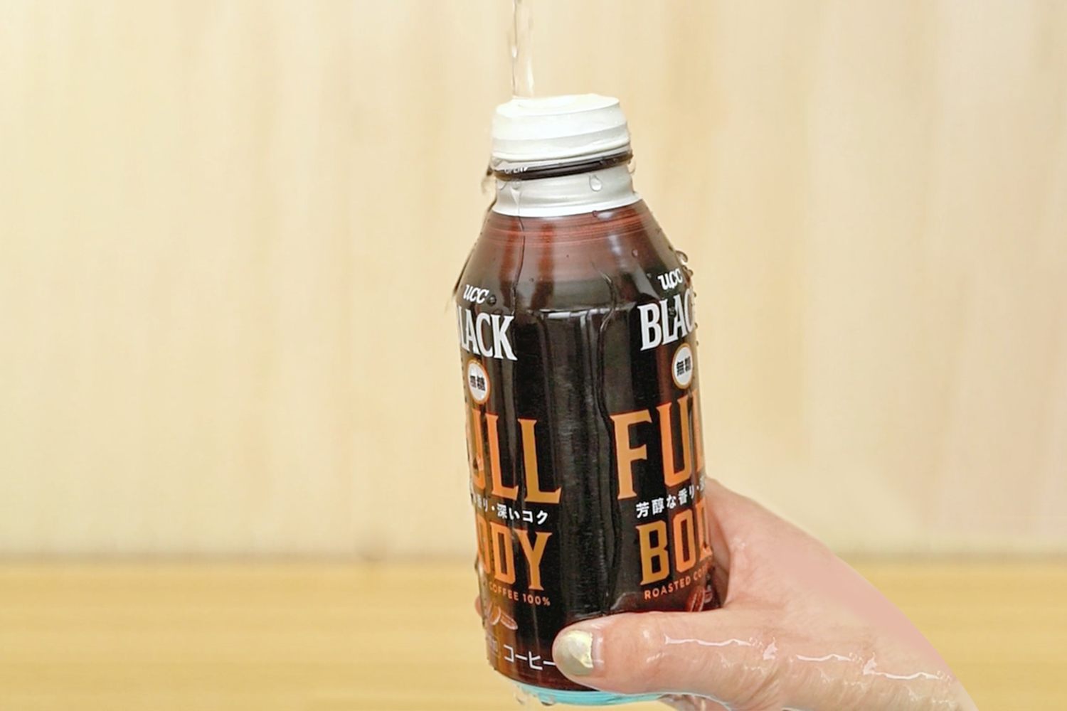 A hand holds a can of Boss Black Full Body coffee under a running tap, against a light brown background.