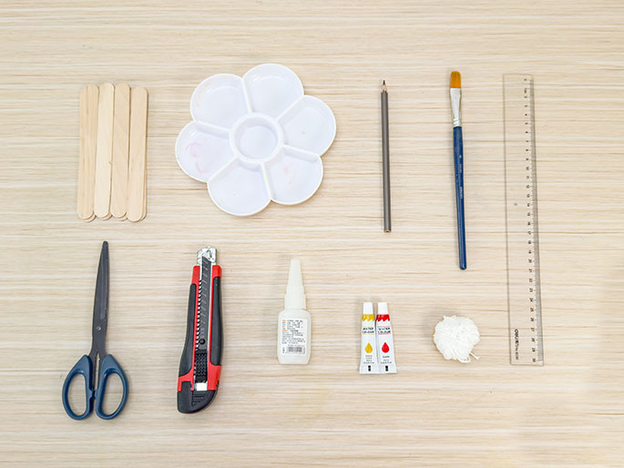 An overhead flatlay shot displays an assortment of craft supplies arranged neatly on a light wooden surface. The items include a bundle of wooden craft sticks, a flower-shaped paint palette, a pencil, a paintbrush, a ruler, scissors, a utility knife, a bottle of glue, two small tubes of paint (red and yellow), and a ball of white yarn or string. The arrangement suggests preparation for a craft project.