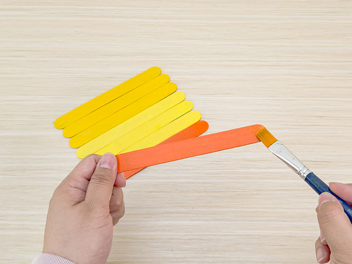 Two hands work on a craft project involving wooden craft sticks on a light wooden surface. One hand holds an orange-painted craft stick, while the other hand uses a paintbrush to apply more orange paint to the end of the stick. In the background, several other craft sticks are visible, painted in shades of yellow, suggesting a gradient effect is being created.