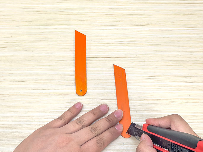 Two hands are working with orange craft sticks on a light wooden surface. One hand holds a stick while the other uses a red and black utility knife to make a precise cut. Another stick, already cut at an angle, lies to the left, indicating steps in a crafting process.