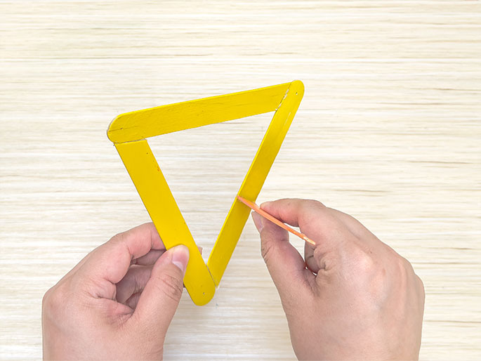 Against a light wooden background, two hands manipulate a triangle constructed from yellow craft sticks. One hand holds the triangle while the other uses a wooden skewer or toothpick to apply glue or some other substance to one of the sticks. This suggests a stage in a craft project where the pieces are being assembled.