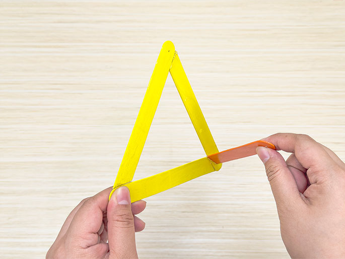 Two hands are assembling a craft project on a light wood background. One hand holds a triangle made of yellow craft sticks, while the other hand positions a smaller, orange craft stick against the base of the triangle. This image appears to capture a moment during the construction of a craft item.