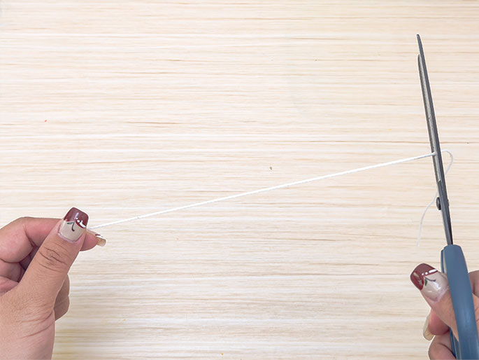 Two hands are shown cutting a length of white string or yarn with scissors against a light wooden background. One hand holds the string taut, while the other hand operates the scissors. The image suggests a simple action of cutting string for a craft or other activity.