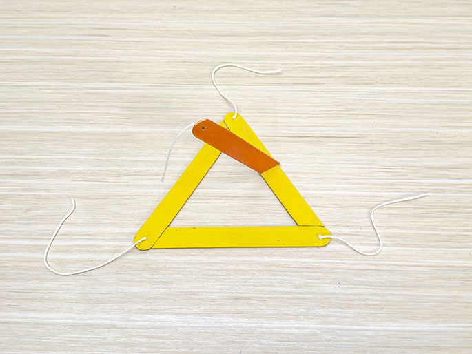 A simple craft project is displayed on a light wooden surface. A triangle formed from yellow craft sticks has a smaller orange stick attached to its top point. Pieces of white string are tied to the two lower corners of the yellow triangle, suggesting the next steps in assembling a hanging mobile or similar object.
