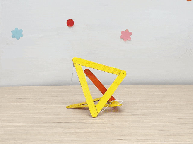 A small, kinetic sculpture made of craft sticks and string rests on a light wood table. The sculpture appears to be in motion, or mid-tumble. It's primarily constructed of yellow sticks, with an orange stick suspended within a triangular frame. White string connects various parts of the sculpture. The background is a plain white wall with small, pastel-colored flower and circle shapes.