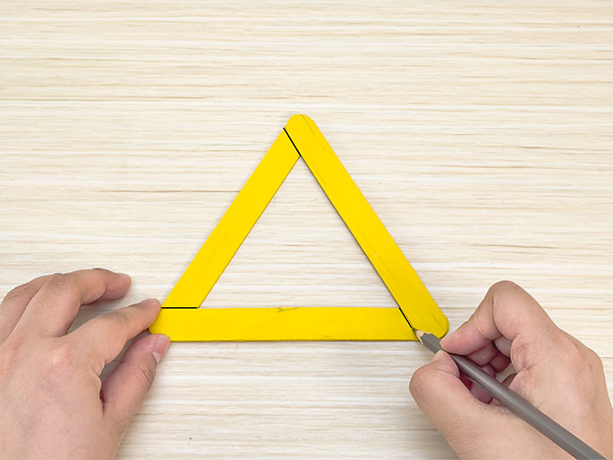 On a light wooden surface, two hands manipulate yellow craft sticks arranged in a triangle. One hand holds the triangle while the other hand uses a pencil to mark lines near the corners of the bottom craft stick, seemingly preparing to cut or modify it.