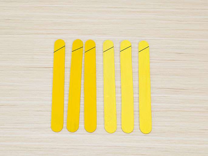 Six yellow craft sticks are arranged vertically on a light wooden background. Each stick has a small diagonal line marked near its rounded top end. This suggests they are being prepared for a craft project and will be cut or shaped along these marked lines.