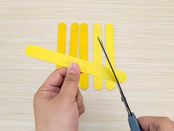 Two hands are positioned above a light wooden surface working with yellow craft sticks. One hand holds a single craft stick horizontally, while the other hand uses scissors to cut the end of that stick. Several more craft sticks stand vertically in the background.