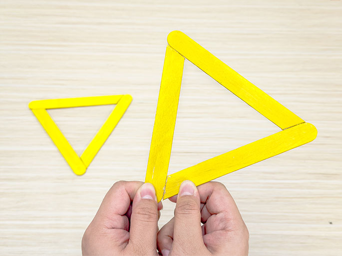 Two hands hold a large triangle made of yellow craft sticks against a light wooden background. A smaller, similarly constructed triangle is visible to the left, suggesting a progression in a craft project.