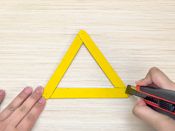 On a light wood-grain background, two hands are shown constructing a triangle from yellow craft sticks. One hand holds the triangle steady, while the other hand uses a red and black utility knife to score or cut one of the sticks.