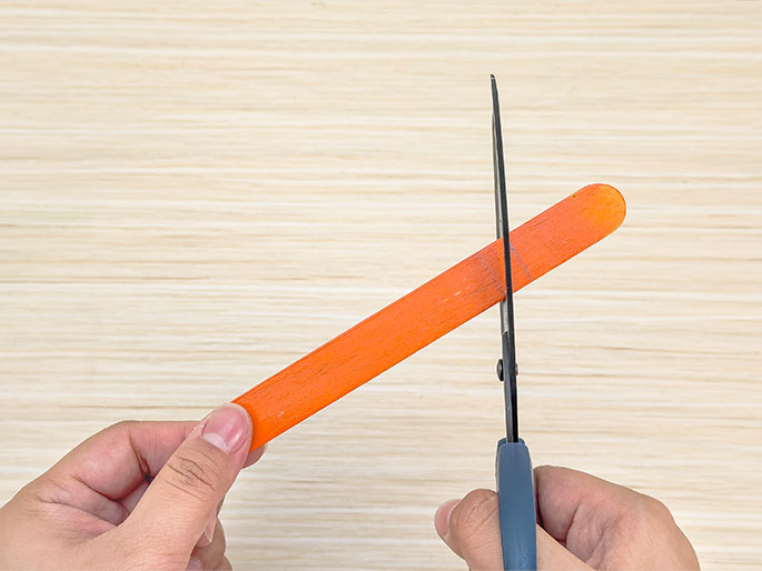 Two hands are shown cutting an orange craft stick with scissors. The craft stick is held diagonally across the image against a light wooden background, and the scissors are positioned to cut across it.