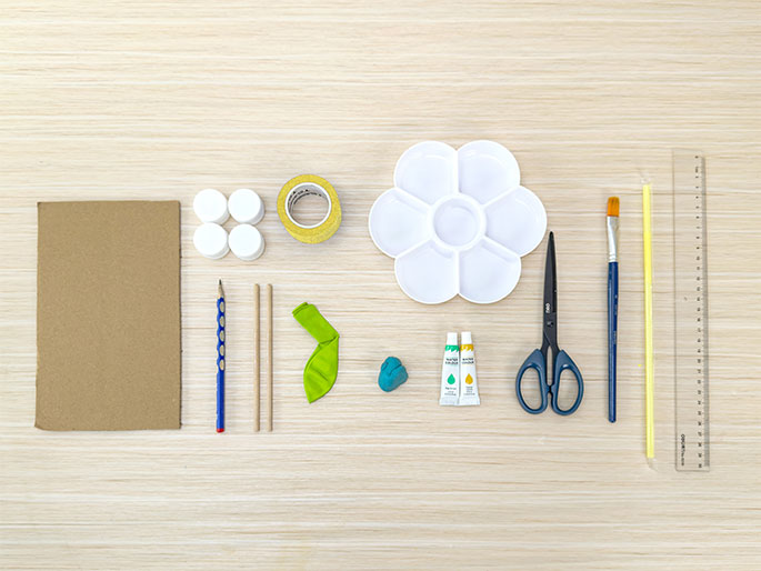 An assortment of craft supplies lies neatly arranged on a light woodgrain surface, viewed from directly above. The items include a piece of brown cardboard, four white plastic bottle caps, a roll of gold glitter tape, a blue pencil, two thin wooden dowels, a deflated lime green balloon, a small lump of teal modeling clay, two small tubes of paint (yellow and teal), a pair of blue scissors, a blue paintbrush, a yellow straw or stick, and a clear plastic ruler. This flat lay suggests materials gathered for a children's craft project.