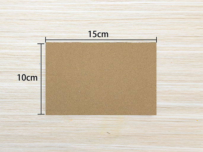 A rectangular piece of light brown cardboard sits on a light-colored wooden surface. Black lines and text indicate its dimensions: 15cm wide and 10cm tall. The image is a simple, straight-on view of the cardboard piece, emphasizing its size and plain surface.