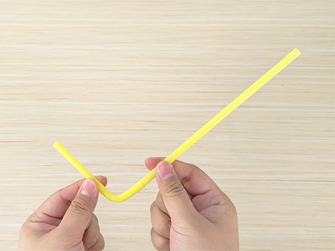 Two hands hold a flexible yellow drinking straw bent into a shallow V shape against a light woodgrain background. The bendable portion of the straw is located near the bottom of the V. The hands gently grip the straw on either side of the bend.