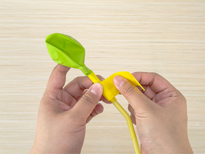 Two hands are shown attaching a deflated lime green balloon to a yellow straw against a light woodgrain background. The neck of the balloon is stretched over the end of the straw. A piece of yellow tape is being wrapped around the connection point to secure the balloon to the straw. One hand holds the balloon and straw while the other hand adjusts the tape.